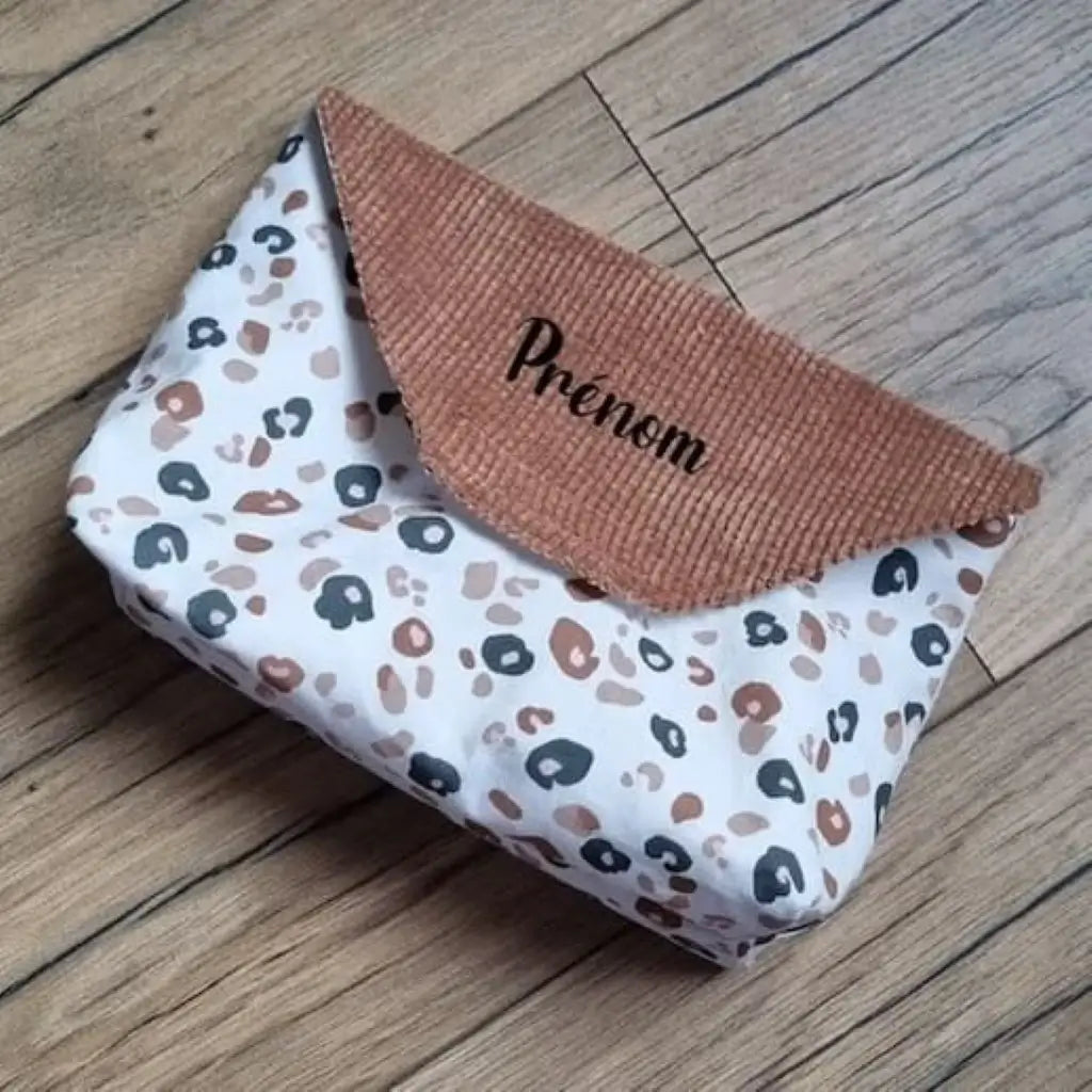 Petite pochette imperméable cousu dans un tissu en coton imprimé au motif léopard et dotée d'un rabat en velours côtelé marron sur lequel on peut floquer un prénom.