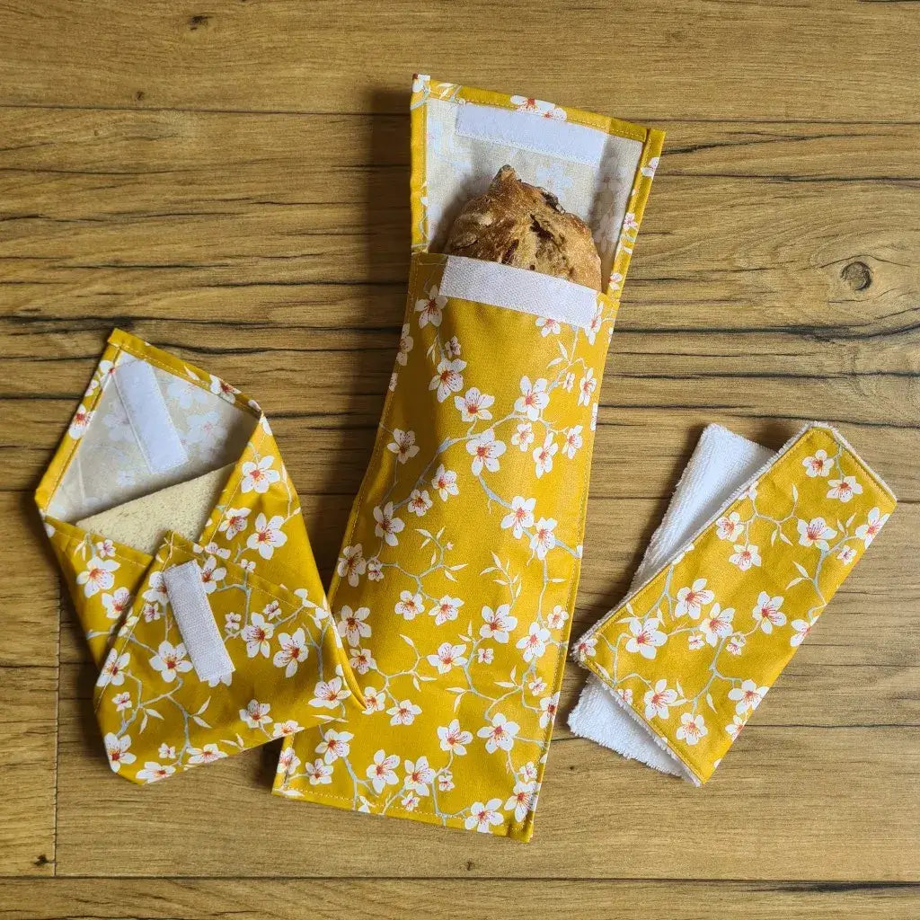 Kit pochettes à sandwichs lavables et réutilisables en coton enduit au motif fleurs jaunes comprenant une pochette au format pain de mie, une pochette au format baguette et un essuie-tout lavable avec une face en éponge de bambou blanche.