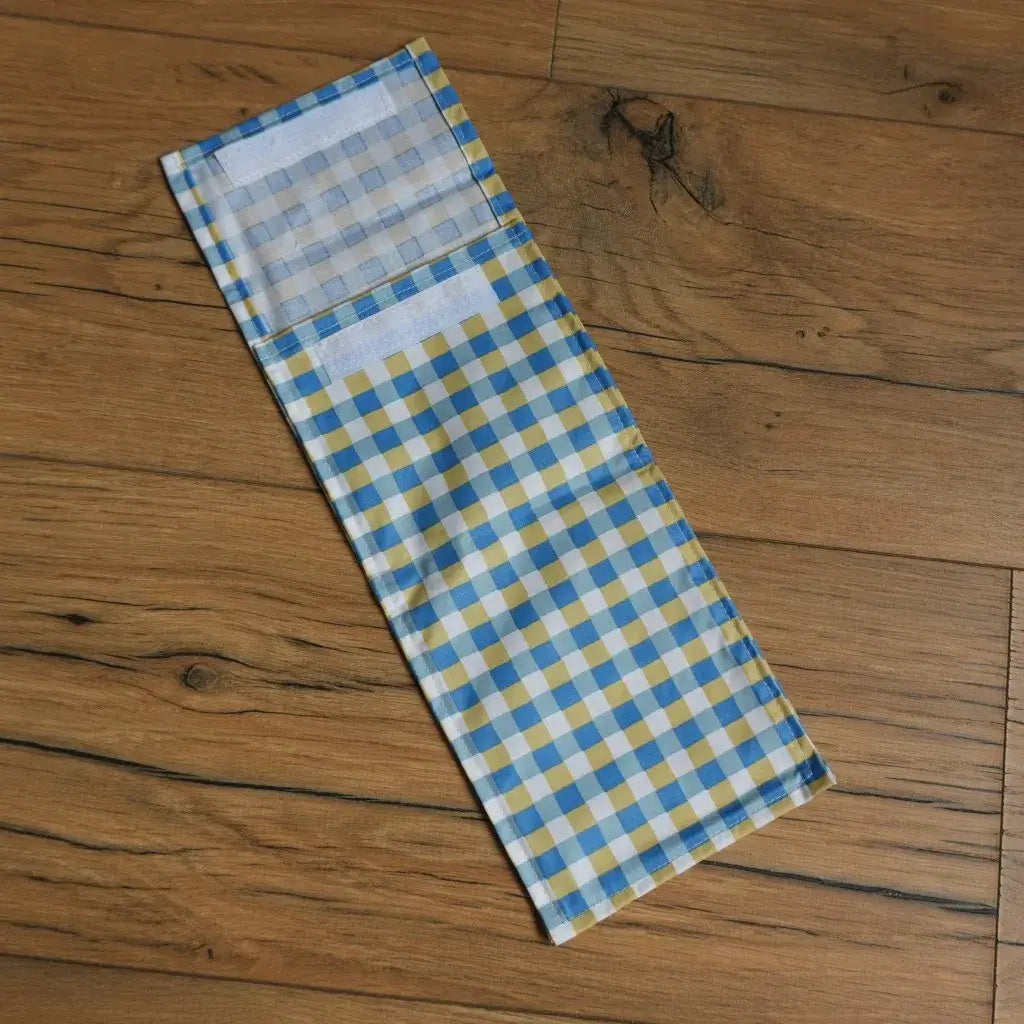 Pochette à sandwich format demi-baguette en tissu enduit à motif carreaux bleus et jaunes, fermée par bande velcro, idéale pour transporter un encas ou un sandwich de manière écologique et réutilisable. Alternative zéro déchet aux emballages jetables pour le déjeuner à l’école ou au bureau.