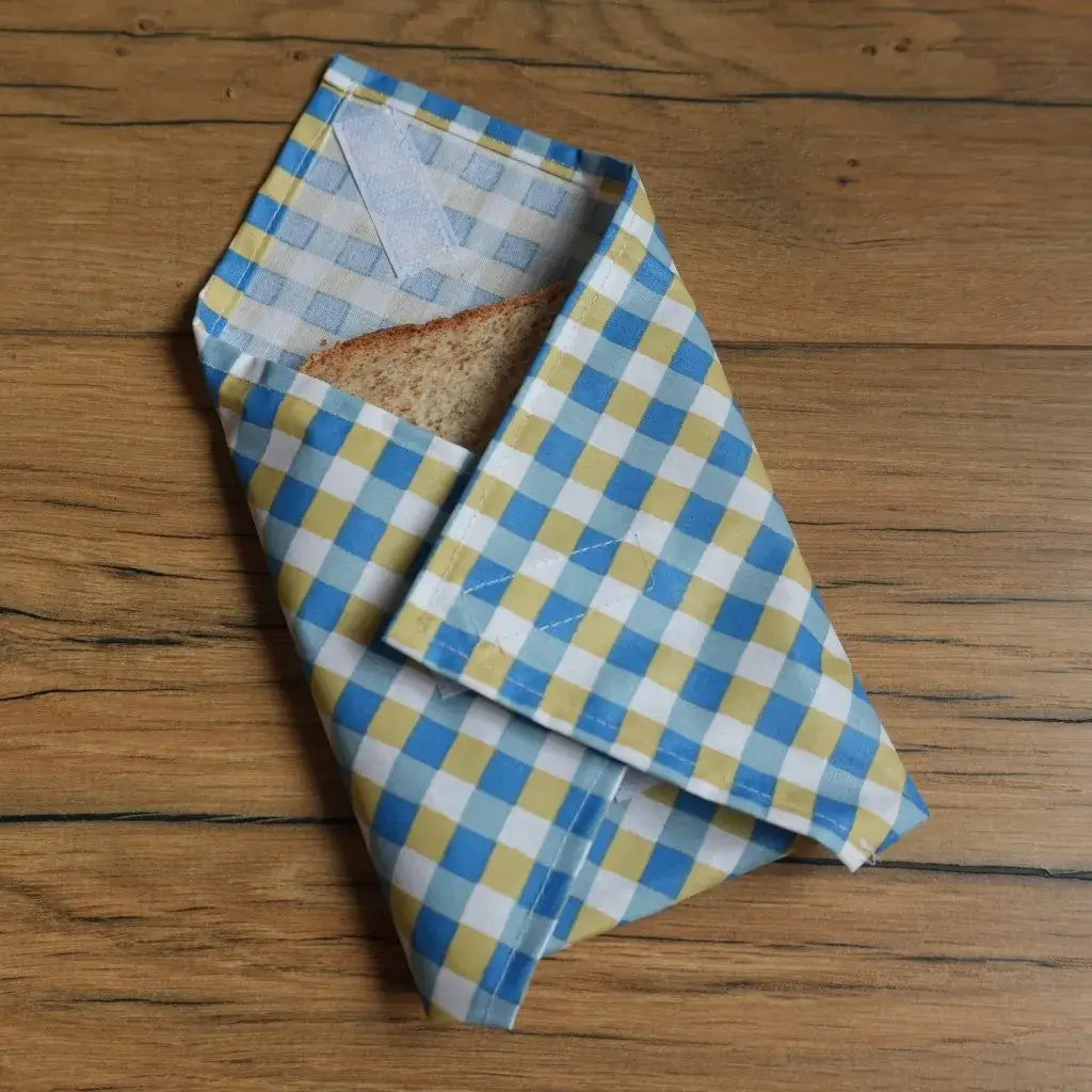 Pochette à sandwich format pain de mie en tissu enduit à carreaux bleus et jaunes, fermée par scratch, contenant une tranche de pain, idéale pour transporter un encas de façon écologique et réutilisable. Emballage alimentaire zéro déchet lavable, parfait pour l’école, le bureau ou les pique-niques.