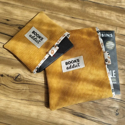 Deux pochettes à liseuse ou livre en velours côtelé jaune disposées l'une sur l'autre. Les deux pochettes comportent la même étiquette en tissu cousue en leur contre et comportant l'inscription "Books addict". Celle du dessus est de petit format pour une liseuse type Kindle ou un livre de poche, celle du dessous est de grand format pour un livre broché.
