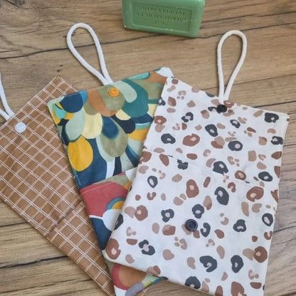 Trois pochettes à savon double compartiment en coton enduit de divers motifs sont disposées en éventail les uns à côté des autres. Ces pochettes disposent d'une cordelette en coton blanc leur permettant d'être suspendues à l'intérieur de la douche. A l'arrière plan, on distingue un savon vert à l'huile d'olive posé juste à côté des pochettes à savon.