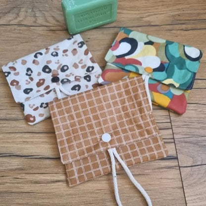 Trois pochettes à savon double compartiment en coton enduit de divers motifs sont disposées en vrac au sol sur un parquet en chêne. Ces pochettes disposent d'une cordelette en coton blanc leur permettant d'être suspendues à l'intérieur de la douche. A l'arrière plan, on distingue un savon vert à l'huile d'olive posé juste à côté des pochettes à savon.