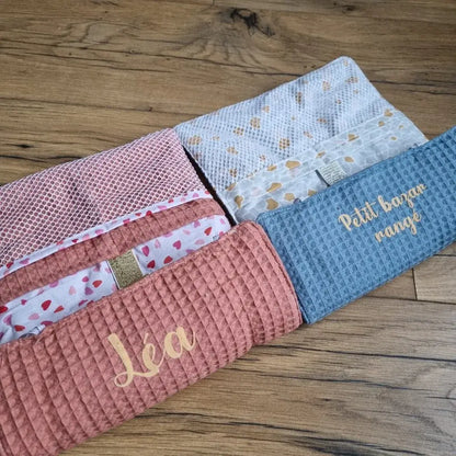Duo de pochettes de rangement multi-usages en nid d'abeille à rouler et personnalisées. Celle de gauche est de coloris marsala, doublée avec un tissu imprimé à motif cœurs, et est personnalisée avec le prénom "Léa". Celle de droite est de coloris bleu denim, doublée avec un tissu imprimé à motifs Argeo et est personnalisée avec l'inscription "Petit bazar rangé".