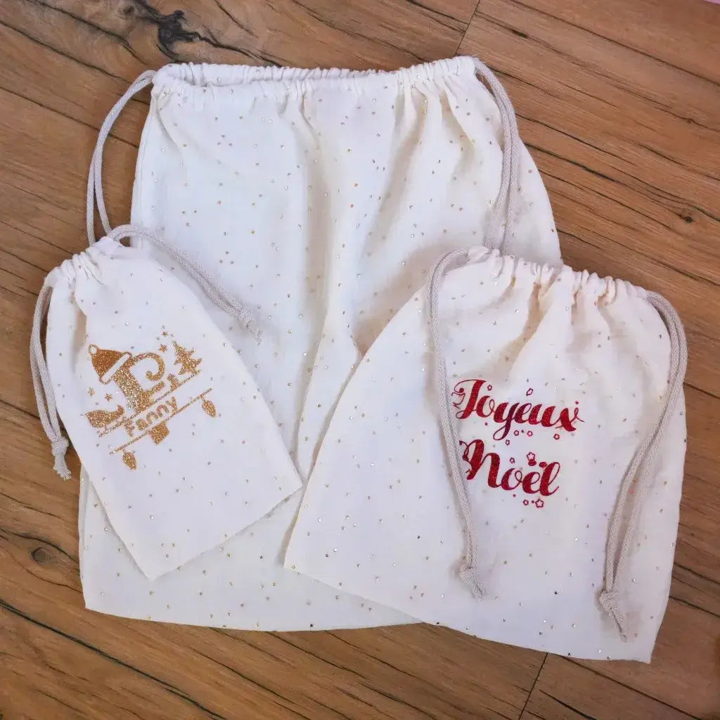 Ensemble de pochons de Noël en double gaze de coton crème à pois dorés, composé de trois tailles différentes. Le plus petit est personnalisé au prénom “Fanny” avec un motif pailleté doré, le moyen porte l’inscription “Joyeux Noël” en rouge scintillant, et le grand reste uni. Ces pochons faits main sont parfaits pour emballer des cadeaux de façon écologique et élégante, tout en apportant une touche artisanale aux fêtes.