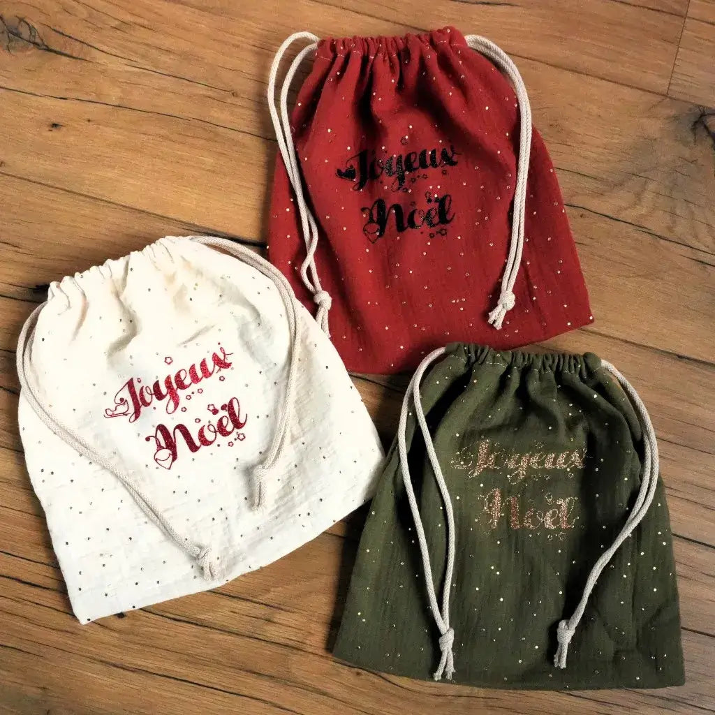 Trio de pochons de Noël en double gaze de coton, déclinés en rouge, vert sapin et écru, chacun décoré de pois dorés et du message “Joyeux Noël” en flocage scintillant. Ces pochettes artisanales à cordon sont idéales pour un emballage cadeau réutilisable et écologique. Leur style chaleureux et festif apporte une touche authentique et raffinée à la décoration de Noël ou au pied du sapin.