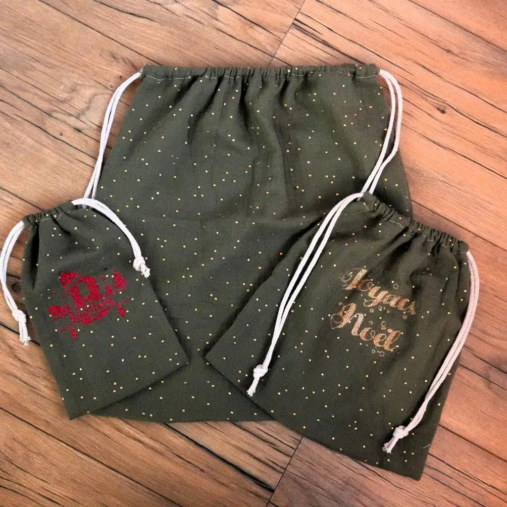 Trio de pochons de Noël en double gaze de coton vert sapin, parsemés de pois dorés pour un effet festif. Le petit modèle est personnalisé au prénom “Baptiste” avec un flocage rouge pailleté, le moyen affiche “Joyeux Noël” en doré, et le grand reste uni. Ces pochons à cordon, faits main, offrent une alternative durable et élégante au papier cadeau, parfaits pour sublimer les présents sous le sapin.