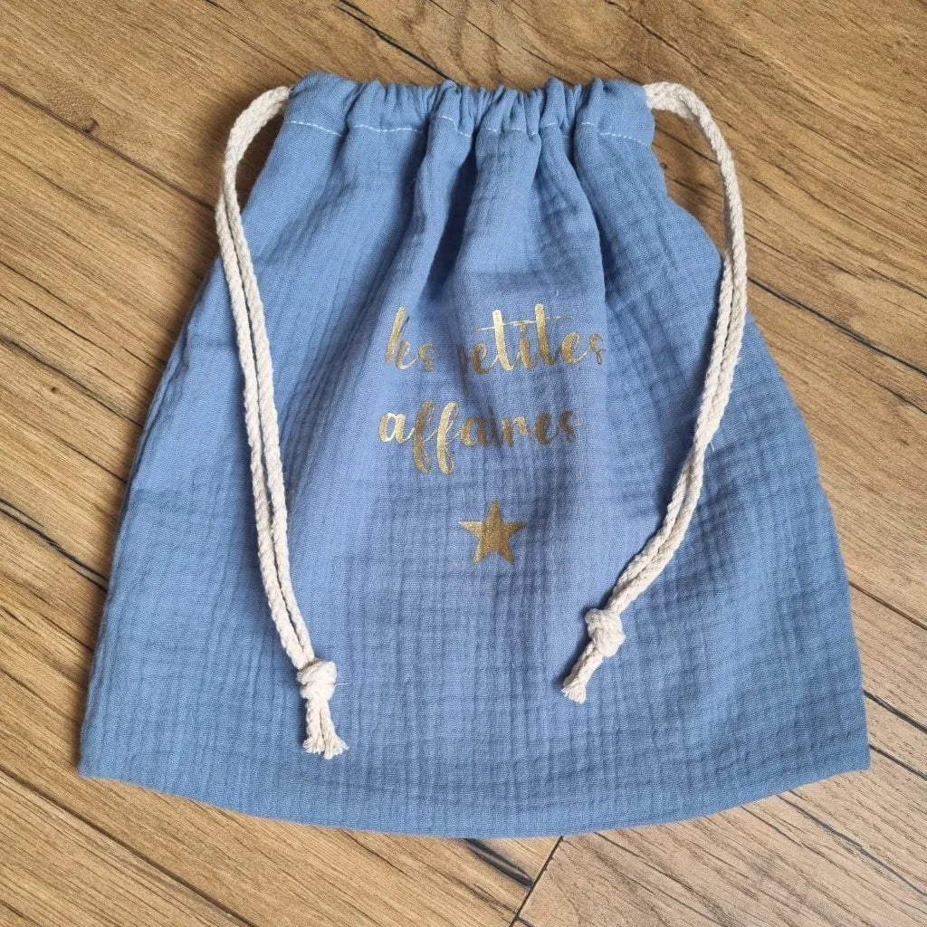 Pochon en tissu double gaze de coton de coloris bleu jeans. Ce pochon se ferme grâce à deux petites cordelettes en coton blanc. Sur ce pochon est floquée une inscription personnalisée "Mes petites affaires" en écriture dorée avec une petite étoile dorée également.