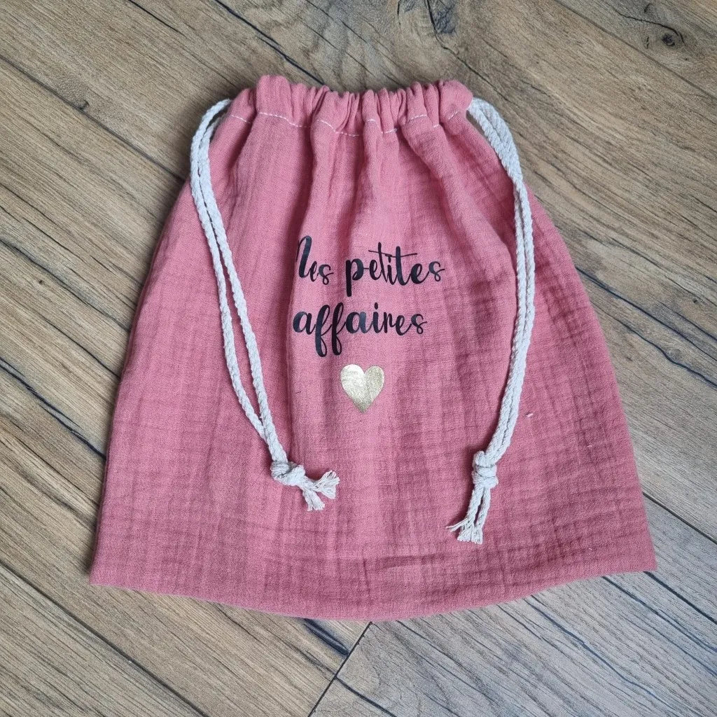 Pochon en tissu double gaze de coton de coloris marsala. Ce pochon se ferme grâce à deux petites cordelettes en coton blanc. Sur ce pochon est floquée une inscription personnalisée "Mes petites affaires" en écriture noire avec un petit cœur doré en-dessous.