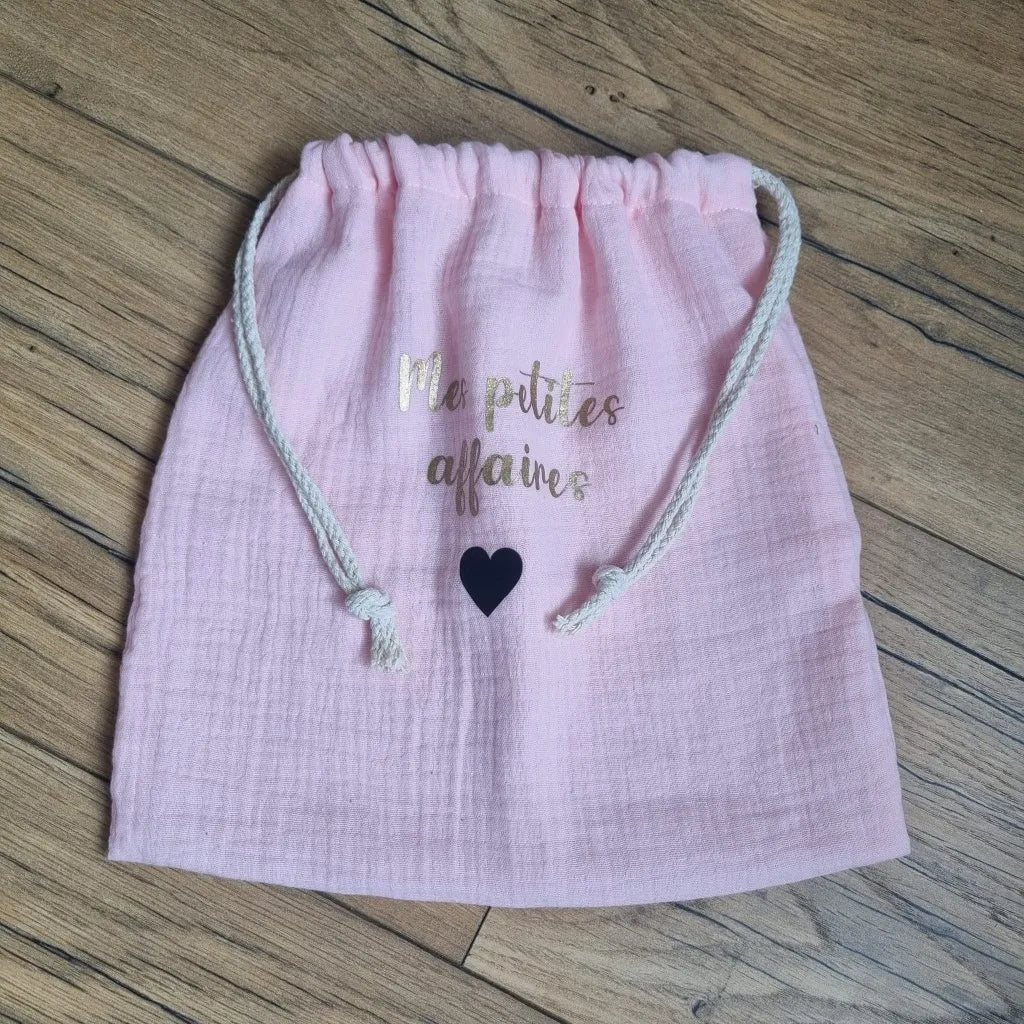 Pochon en tissu double gaze de coton de coloris rose pâle. Ce pochon se ferme grâce à deux petites cordelettes en coton blanc. Sur ce pochon est floquée une inscription personnalisée "Mes petites affaires" en écriture dorée avec un petit cœur en noir en-dessous.