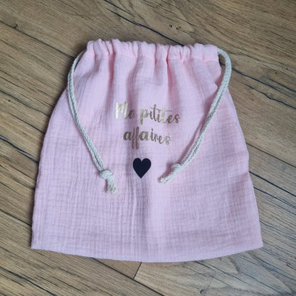 Pochon en tissu double gaze de coton de coloris rose pâle. Ce pochon se ferme grâce à deux petites cordelettes en coton blanc. Sur ce pochon est floquée une inscription personnalisée "Mes petites affaires" en écriture dorée avec un petit cœur en noir en-dessous.