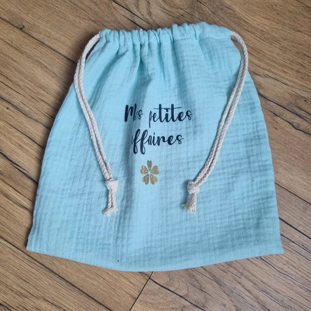 Pochon en tissu double gaze de coton de coloris vert menthe. Ce pochon se ferme grâce à deux petites cordelettes en coton blanc. Sur ce pochon est floquée une inscription personnalisée "Mes petites affaires" en écriture noire avec une petite fleur dorée en-dessous.