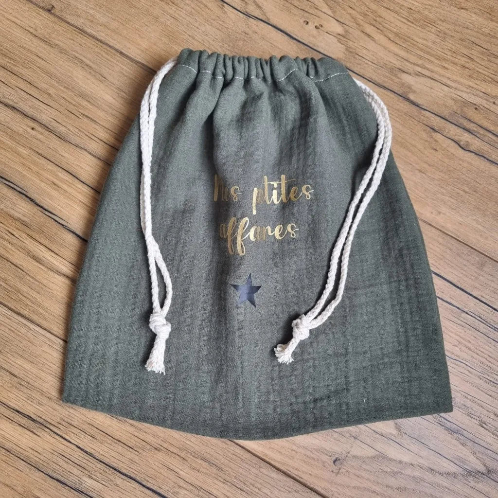 Pochon en tissu double gaze de coton de coloris vert. Ce pochon se ferme grâce à deux petites cordelettes en coton blanc. Sur ce pochon est floquée une inscription personnalisée "Mes petites affaires" en écriture dorée avec une petite étoile noire en dessous.