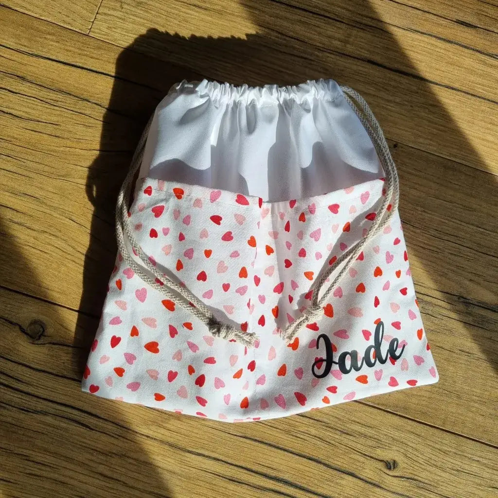 Pochon personnalisé pour enfant en tissu enduit blanc imprimé de petits cœurs rouges et roses, avec cordon de serrage beige et prénom Jade floqué en noir. Sac pratique et léger, idéal pour la crèche, la maternelle, le sport ou la piscine. Accessoire unique et artisanal qui allie fonctionnalité et touche tendre, parfait comme cadeau personnalisé.