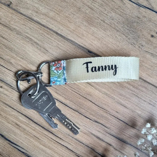 Porte-clés dragonne personnalisé avec le prénom 'Fanny', en sangle écrue et tissu fleuri, accroché à un trousseau de clés. Accessoire pratique et original à offrir ou à s’offrir.
