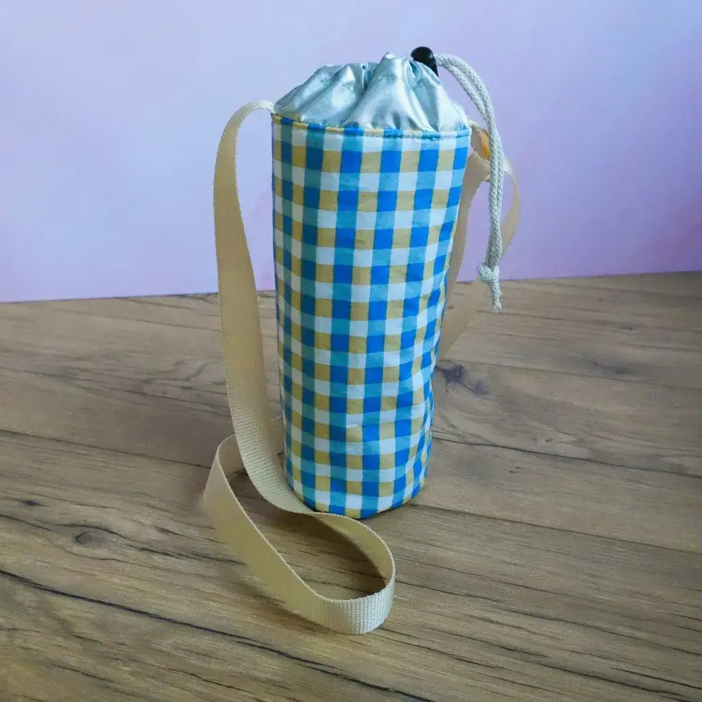 Porte-gourde isotherme à motif carreaux vichy bleus et jaunes, bandoulière beige, sur une table en bois.