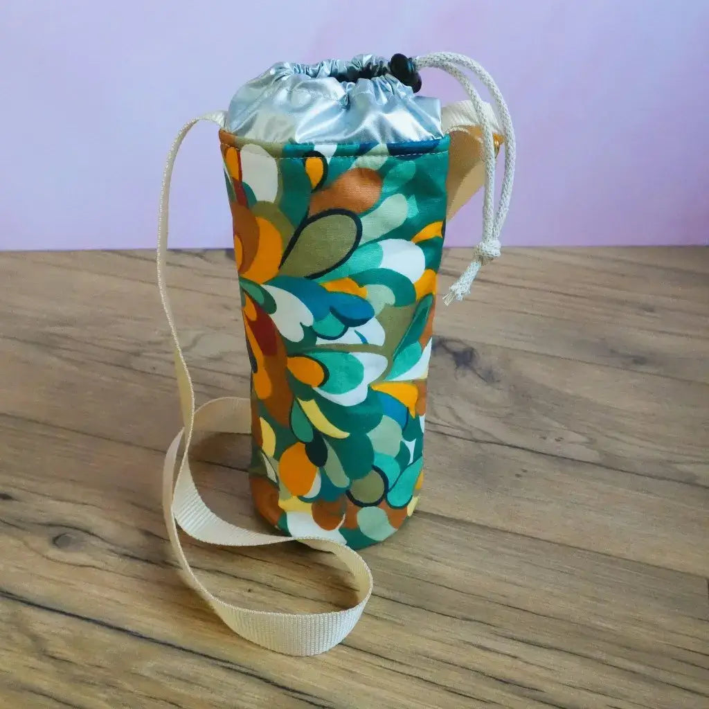 Porte-gourde isotherme en coton enduit motif splash aux formes abstraites orange et vert canard, avec bandoulière beige.