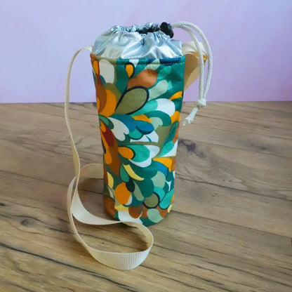 Porte-gourde isotherme en coton enduit motif splash aux formes abstraites orange et vert canard, avec bandoulière beige.