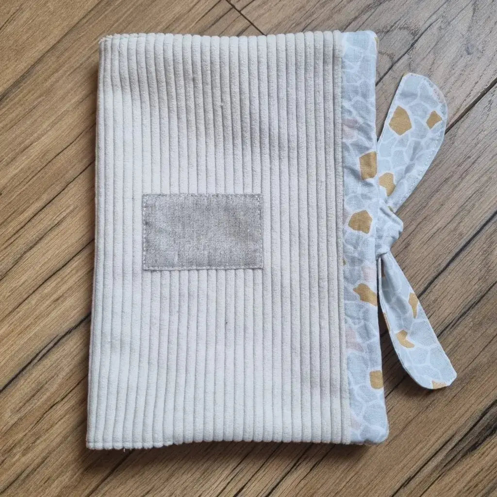 Protège carnet de santé en velours côtelé de coloris beige. Ce protège carnet de santé se ferme à l'aide d'un ruban en tissu imprimé au motif argéo. Il est personnalisable avec le prénom de l'enfant floqué sur une petite étiquette en tissu.