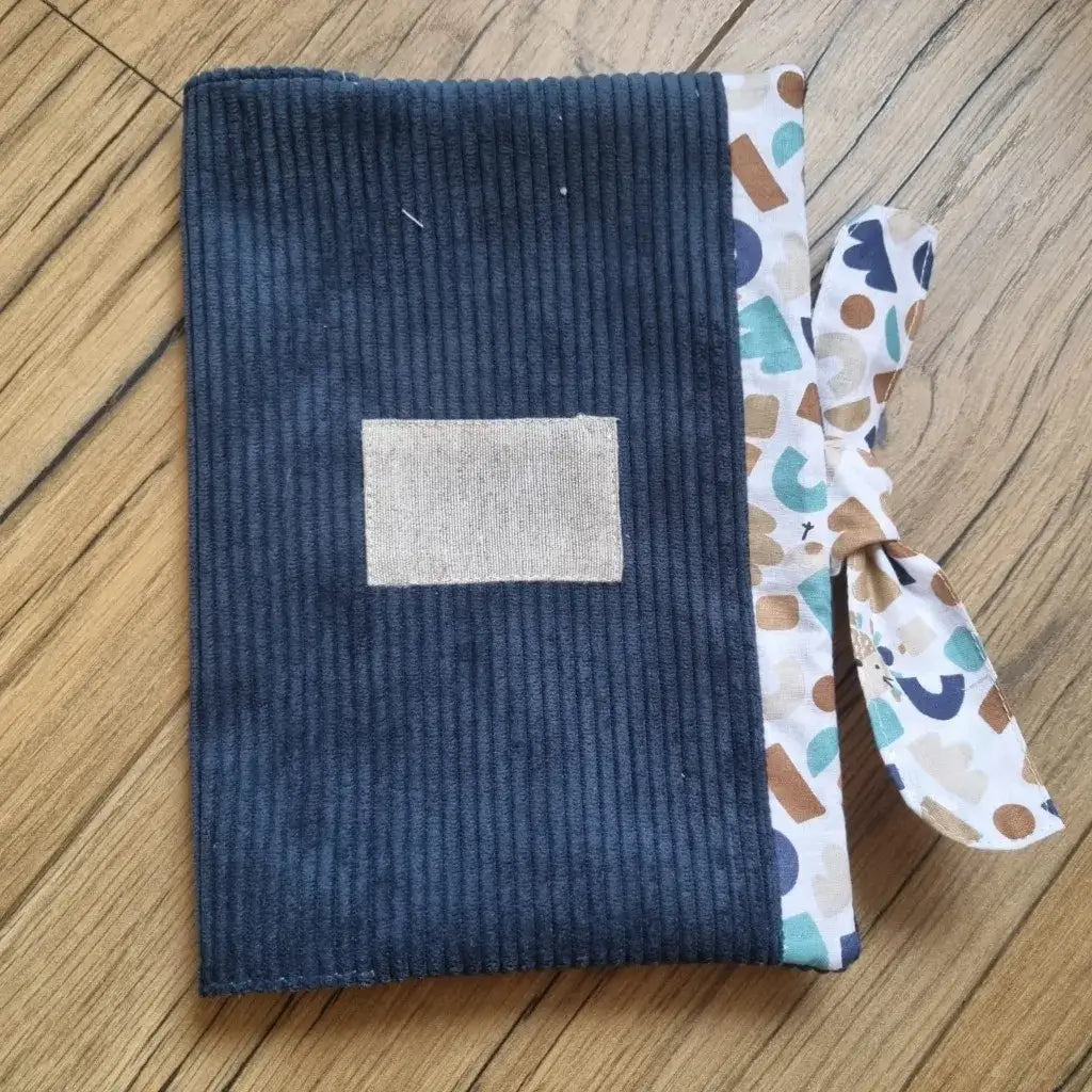 Protège carnet de santé en velours côtelé de coloris bleu foncé. Ce protège carnet de santé se ferme à l'aide d'un ruban en tissu imprimé au motif ptiwi. Il est personnalisable avec le prénom de l'enfant floqué sur une petite étiquette en tissu.