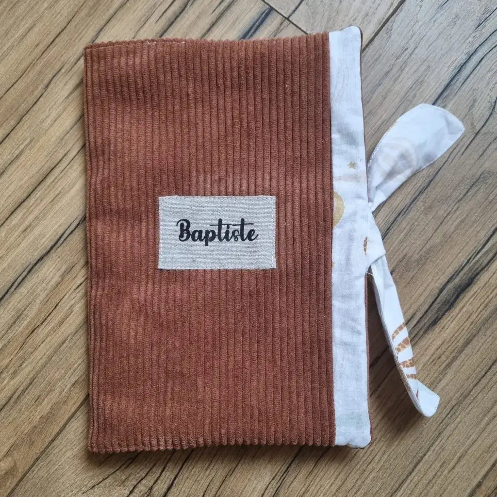 Protège carnet de santé en velours côtelé de coloris marron. Ce protège carnet de santé se ferme à l'aide d'un ruban en tissu imprimé au motif montgolfière. Il est ici personnalisé avec le prénom "Baptiste" floqué sur une petite étiquette en tissu.