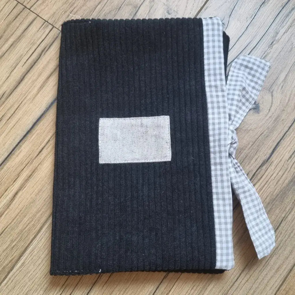 Protège carnet de santé en velours côtelé de coloris noir. Ce protège carnet de santé se ferme à l'aide d'un ruban en tissu imprimé au motif carreaux beiges. Il est personnalisable avec le prénom de l'enfant floqué sur une petite étiquette en tissu.