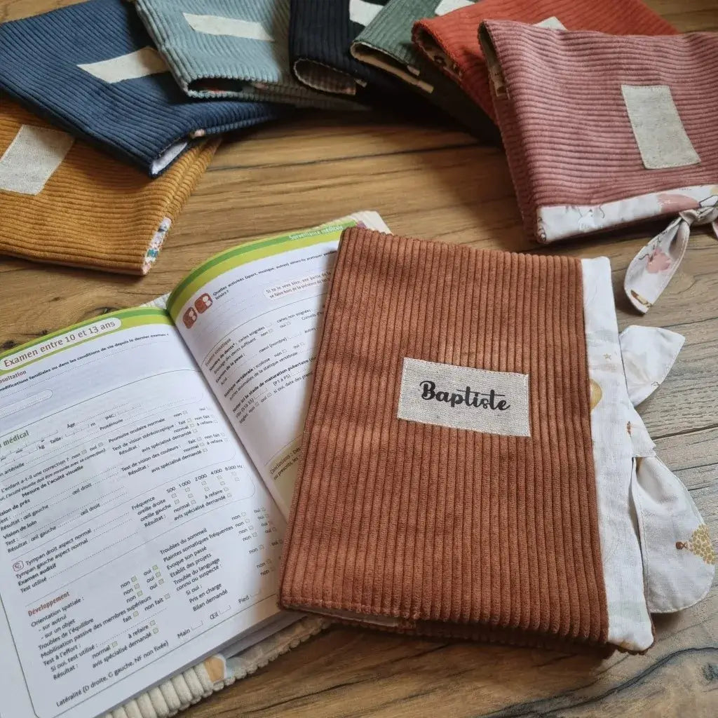 Au premier plan, un protège carnet de santé en velours côtelé marron personnalisé avec le prénom "Baptiste" floqué sur une petite étiquette en tissu cousue en son centre. Ce protège carnet de santé est posé sur un carnet de santé ouvert sur une double page. A l'arrière plan, des protège carnets de santé identiques mais dans divers coloris différents et disposés au sol en éventail.