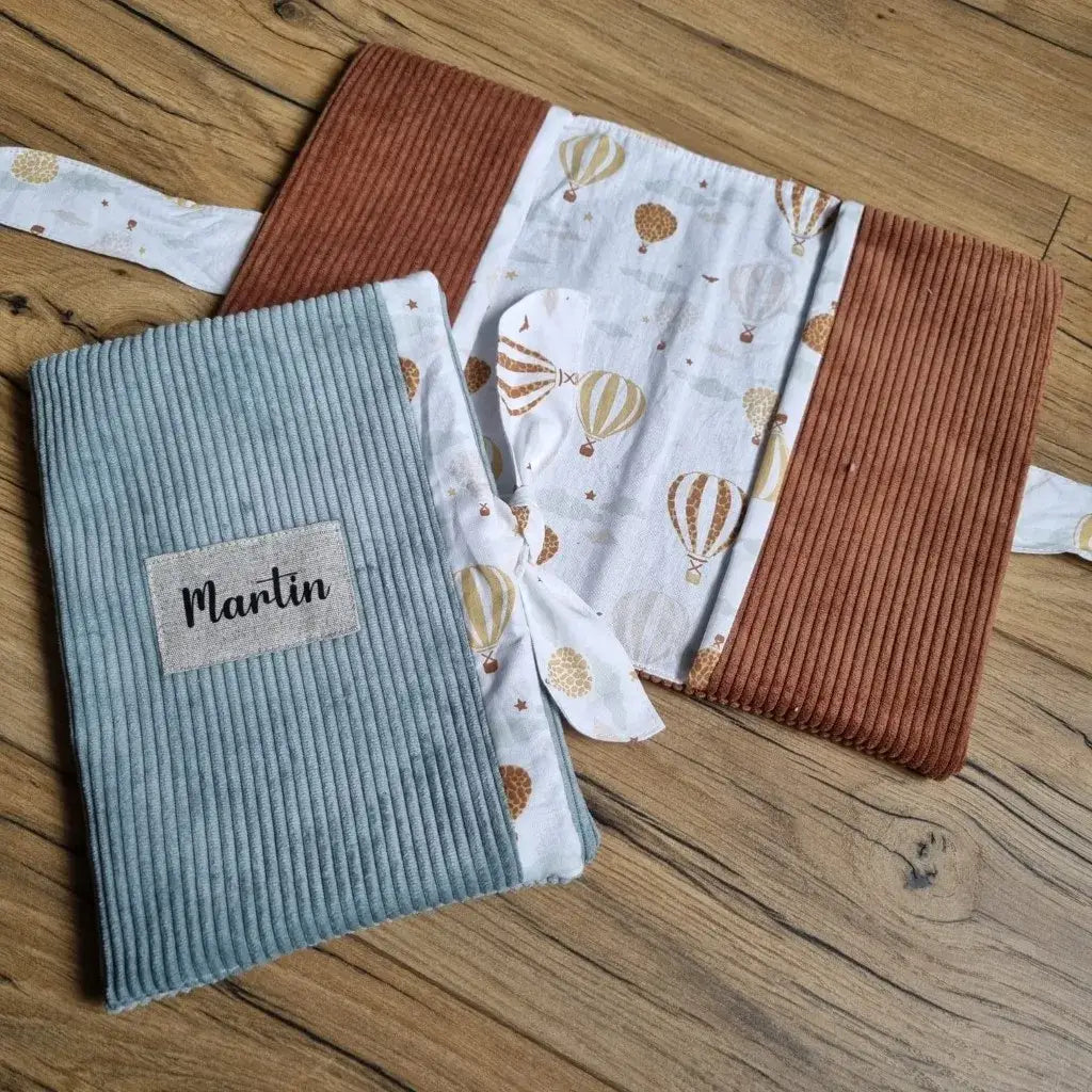 Au premier plan, un protège carnet de santé en velours côtelé de coloris bleu clair. Ce protège carnet de santé est fermé grâce à un ruban en tissu imprimé au motif montgolfières et est personnalisé avec le prénom "Martin" floqué sur une petite étiquette en tissu. A l'arrière plan, le même carnet de santé mais en velours côtelé marron et complètement ouvert.