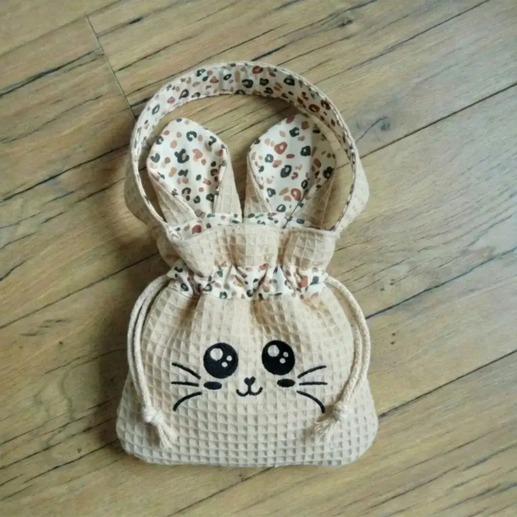 Sac pochon beige naturel en nid d'abeille avec visage Chatoune noir et doublure à motifs léopard, posé sur un parquet.