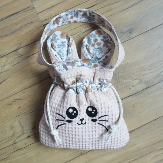 Pochon couleur peau en nid d'abeille avec flocage Chatoune noir, oreilles fleuries et cordon blanc, posé à plat sur un parquet.