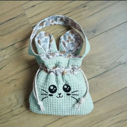 Sac pochon vert menthe en nid d'abeille avec visage Chatoune noir et doublure fleurie assortie, posé sur un parquet.