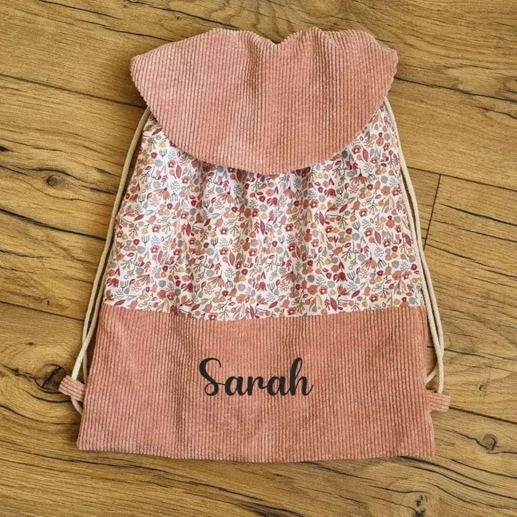 Sac à dos enfant au format pochon avec rabat personnalisé avec le prénom "Sarah". Ce sac est cousu avec un tissu imprimé au motif Liberty. Sa base et son rabat sont en velours côtelé de coloris rose.