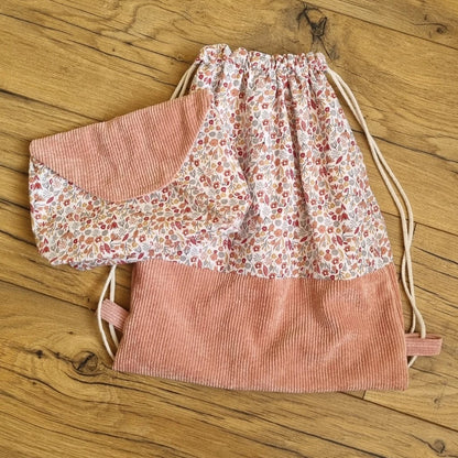 Sac à dos format pochon avec une base en velours côtelé rose. Le corps du sac est composé d'un tissu au motif liberty. A gauche de ce sac est disposée une pochette assortie avec le même tissu motif liberty et un rabat en velours côtelé rose.