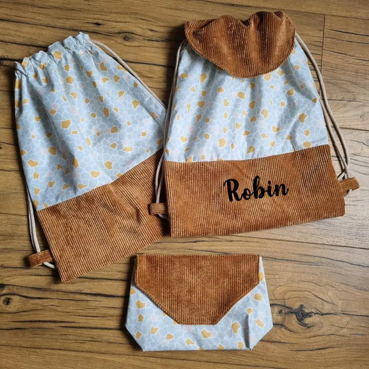 Ensemble de trois accessoires cousus main posés sur un sol en bois : un sac à dos personnalisé au prénom "Robin", un pochon à cordon et une pochette assortie. Chaque pièce est confectionnée en velours côtelé marron et tissu à motifs abstraits bleu clair et moutarde. Idéal pour un cadeau enfant original, une rentrée scolaire ou un usage quotidien pratique et stylé.