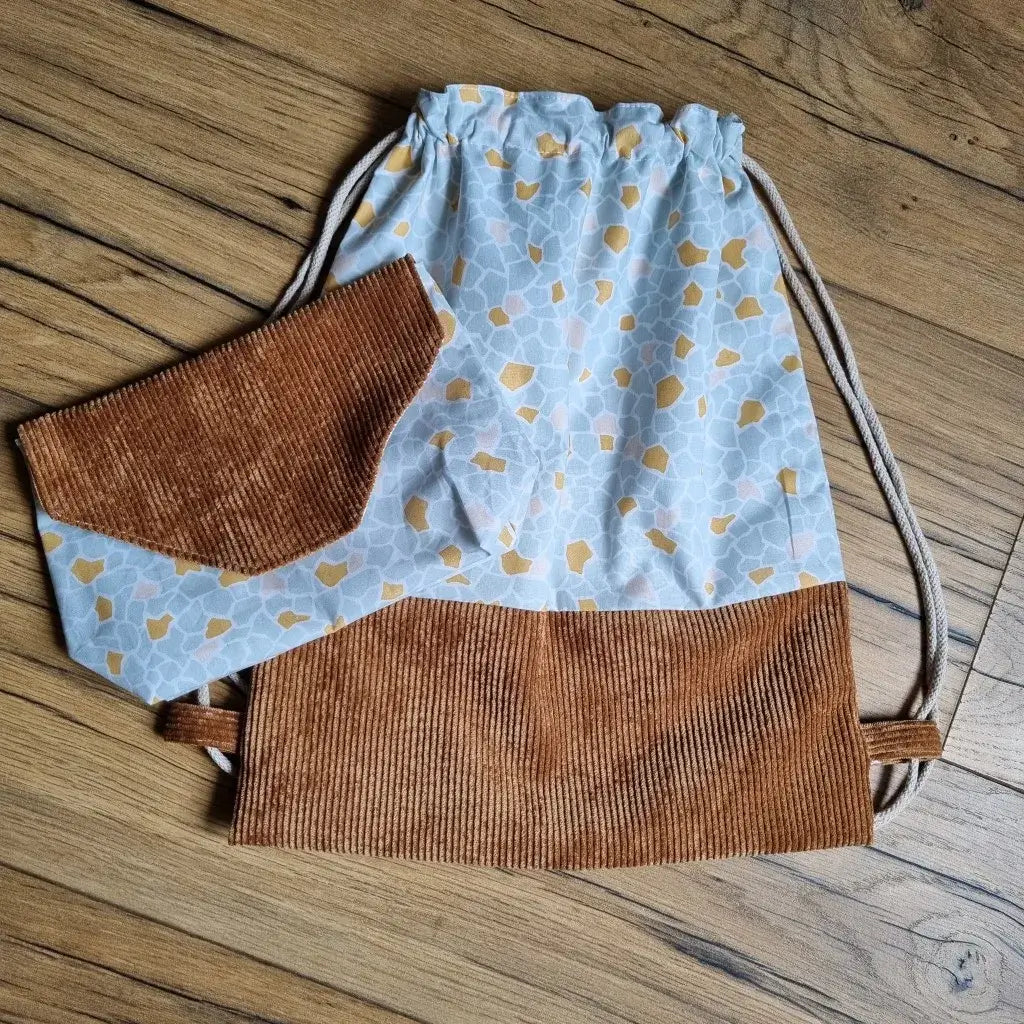 Sac à dos pochon sans rabat et pochette assortie posés sur un sol en bois, réalisés en tissu à motifs bleu clair et moutarde, avec empiècements en velours côtelé marron. Le sac est équipé de cordons beiges pour un port confortable, tandis que la pochette présente un rabat en V inversé. Ensemble fait main pratique et coordonné, parfait pour l’école, la crèche ou en cadeau personnalisé.