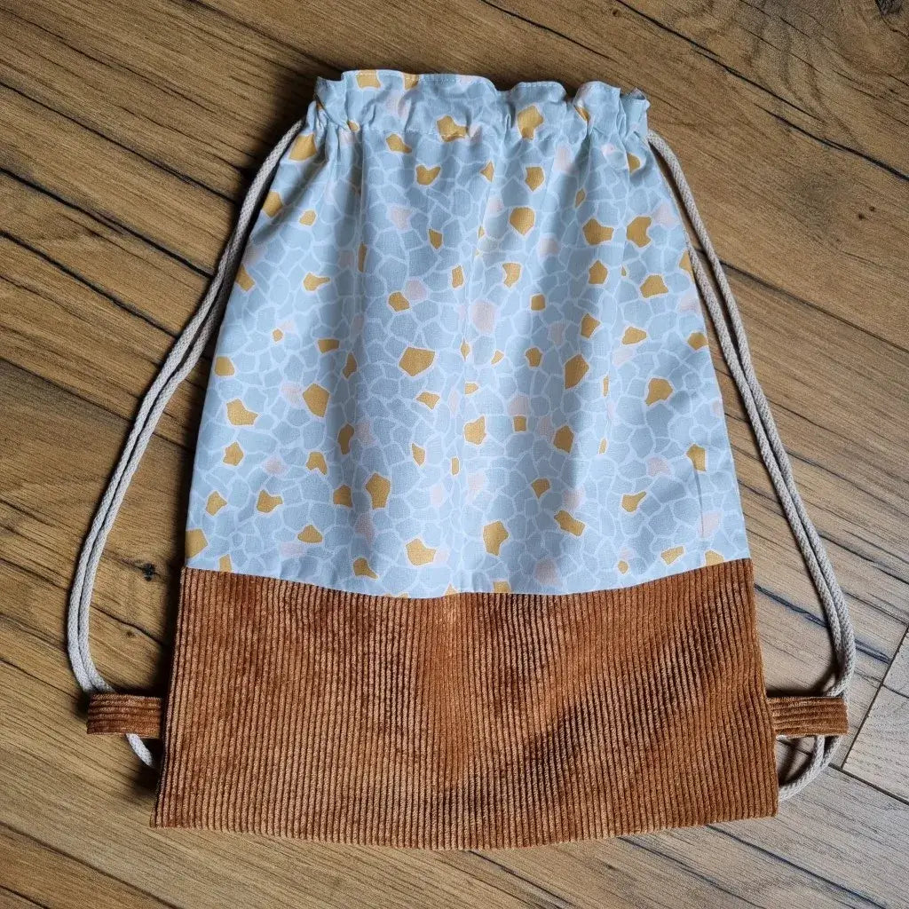 Sac à dos pochon artisanal sans rabat, posé à plat sur un sol en bois, confectionné en tissu imprimé bleu clair à motifs moutarde et rose pâle, avec une base en velours côtelé marron. Fermeture par cordons beiges pour un port confortable sur les épaules. Accessoire pratique, léger et tendance, idéal pour la crèche, l’école ou les activités sportives des enfants.
