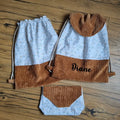Ensemble cousu main composé de deux sacs à dos pochons en tissu floral bleu clair et velours côtelé marron, dont un personnalisé avec le prénom "Diane", accompagné d’une pochette assortie. Fermeture par cordons coulissants en coton naturel. Accessoires pratiques et élégants, parfaits pour l’école, les sorties ou en cadeau unique. Posés sur un sol en bois.