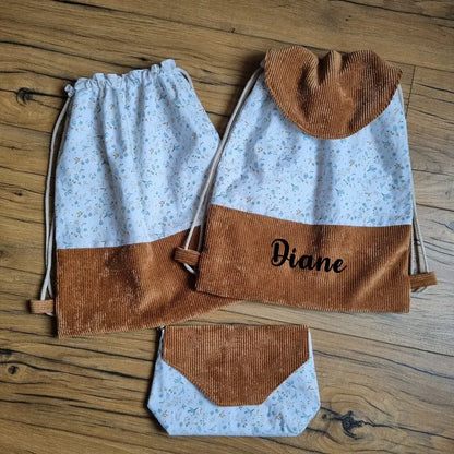 Ensemble cousu main composé de deux sacs à dos pochons en tissu floral bleu clair et velours côtelé marron, dont un personnalisé avec le prénom "Diane", accompagné d’une pochette assortie. Fermeture par cordons coulissants en coton naturel. Accessoires pratiques et élégants, parfaits pour l’école, les sorties ou en cadeau unique. Posés sur un sol en bois.
