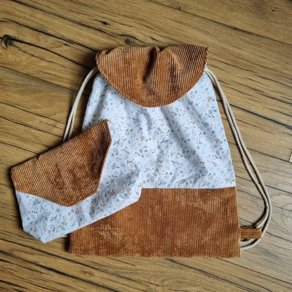 Ensemble cousu main composé d’un sac à dos pochon avec rabat en velours côtelé marron et tissu fleuri bleu clair, accompagné d’une pochette assortie. Fermeture du sac par cordons coulissants en coton naturel. Accessoires pratiques et élégants, parfaits pour une rentrée personnalisée ou un cadeau unique. Posés sur un sol en bois.