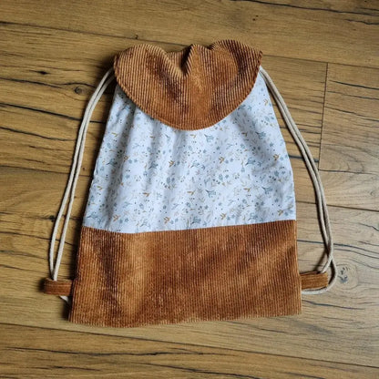 Sac à dos pochon artisanal en tissu coton à motif floral bleu clair et base en velours côtelé marron, avec rabat arrondi assorti. Fermeture par cordons coulissants en coton naturel. Modèle fait main élégant et pratique, idéal pour les enfants, les sorties ou en cadeau personnalisé. Accessoire posé sur un sol en bois.