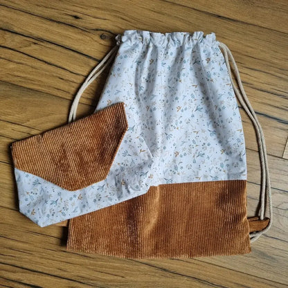 Ensemble cousu main composé d’un sac à dos pochon sans rabat en coton blanc à motif floral bleu et base en velours côtelé marron, accompagné d’une pochette assortie avec rabat. Fermeture du sac par cordons coulissants en coton naturel. Accessoires pratiques, esthétiques et personnalisables, posés sur un sol en bois.