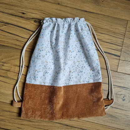 Sac à dos pochon artisanal sans rabat, confectionné en coton blanc à motif floral bleu et ocre, avec base en velours côtelé marron. Fermeture par cordons coulissants en coton naturel. Accessoire fait main pratique, léger et esthétique, idéal pour les enfants, les loisirs ou un cadeau personnalisé. Posé sur un sol en bois.