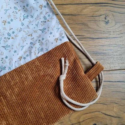 Gros plan sur la base d’un sac à dos pochon artisanal en velours côtelé camel et tissu fleuri bleu sur fond blanc, mettant en évidence la finition soignée, la couture robuste et la cordelette en coton naturel nouée. Accessoire fait main élégant et durable, idéal pour un usage quotidien, posé sur un sol en bois.