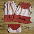 Ensemble d’accessoires cousus main composé de deux sacs à dos pochons en tissu fleuri et velours côtelé terracotta, dont un personnalisé avec le prénom "Cathy", et d’une pochette assortie. Parfait pour l’école, les sorties ou les cadeaux personnalisés. Posés sur un sol en bois. Style artisanal, féminin et tendance.
