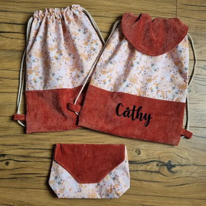 Ensemble d’accessoires cousus main composé de deux sacs à dos pochons en tissu fleuri et velours côtelé terracotta, dont un personnalisé avec le prénom "Cathy", et d’une pochette assortie. Parfait pour l’école, les sorties ou les cadeaux personnalisés. Posés sur un sol en bois. Style artisanal, féminin et tendance.