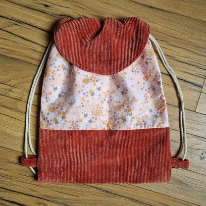 Sac à dos pochon artisanal en tissu fleuri rose et base en velours côtelé terracotta, avec rabat arrondi assorti. Fermeture par cordons coulissants en coton naturel. Idéal pour les enfants ou les sorties décontractées. Modèle fait main, pratique et tendance, posé sur un sol en bois.