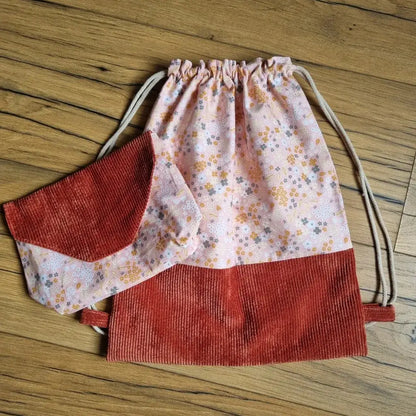 Ensemble artisanal composé d’un sac à dos pochon sans rabat en tissu fleuri rose et velours côtelé terracotta, assorti à une pochette avec rabat coordonné. Fermeture du sac par cordons coulissants. Idéal pour l’école, les sorties ou en cadeau personnalisé. Accessoires faits main, posés sur un sol en bois.