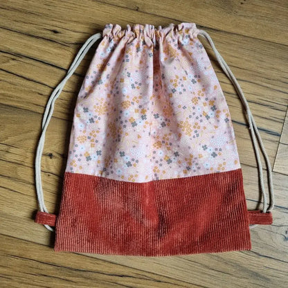 Sac à dos pochon artisanal sans rabat, confectionné en tissu fleuri rose à motifs floraux et base en velours côtelé terracotta, avec fermeture par cordons coulissants en coton naturel. Accessoire fait main pratique et léger, idéal pour l’école, les sorties ou les loisirs, posé sur un sol en bois.