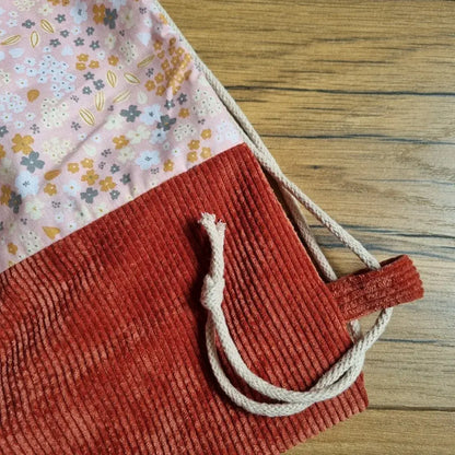 Gros plan sur la base d’un sac à dos pochon fait main en velours côtelé terracotta et tissu fleuri rose, mettant en valeur la finition soignée de la couture et la cordelette épaisse en coton naturel insérée dans une attache latérale. Accessoire artisanal robuste et esthétique, posé sur un sol en bois.