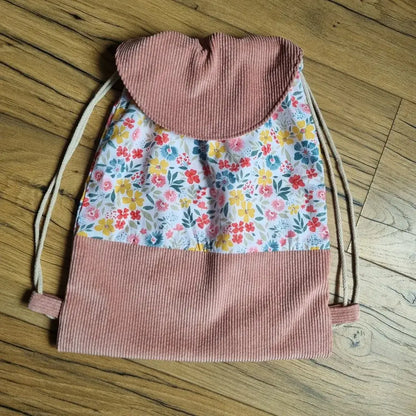 Sac à dos pochon fait main, confectionné en tissu fleuri multicolore avec base et rabat en velours côtelé rose. Fermeture par cordon coulissant beige et rabat arrondi pour un style tendre et champêtre. Idéal pour les enfants, ce sac allie praticité, confort et esthétisme, parfait pour l’école, les sorties ou comme cadeau personnalisé. Présenté sur un fond en bois chaleureux.